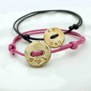 Puede incluir: Dos pulseras con dijes de botón de oro. Un dije dice "Papá" con "XX" arriba. El otro dije dice "B ♥ L".