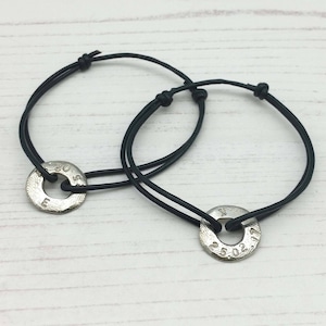May include: Two black leather bracelets with silver metal charms. The charms are round and have engraved text. The text on the charms reads "20.05" and "25.02".