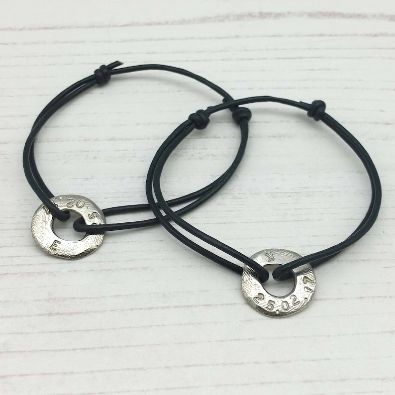 May include: Two black leather bracelets with silver metal charms. The charms are round and have engraved text. The text on the charms reads "20.05" and "25.02".