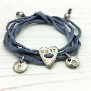 May include: A blue wrap bracelet with a heart-shaped charm engraved with "2011" and two round charms with the letters "T" and "B". The bracelet is made of multiple strands of blue fabric and has silver-colored beads at the ends.