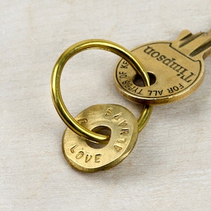 Op de afbeelding: Een gouden sleutelhanger met een sleutel eraan en twee ronde metalen labels. De sleutel is gelabeld "Voor alle soorten sleutels" en "Thompson". Een label zegt "Liefde" en de andere zegt "Altijd".