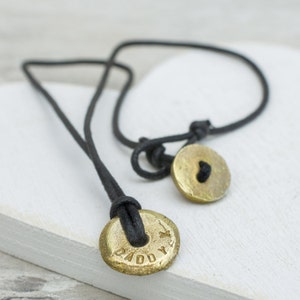 May include: A black cord necklace with two gold round pendants. One pendant has the word "Daddy" engraved on it and the other has a heart engraved on it.