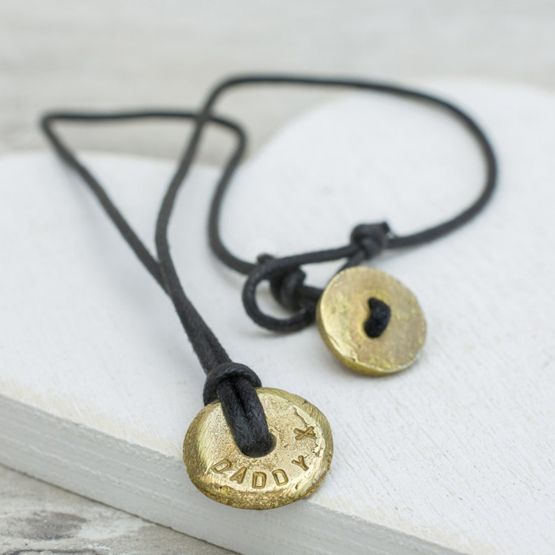 May include: A black cord necklace with two gold round pendants. One pendant has the word "Daddy" engraved on it and the other has a heart engraved on it.