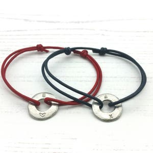 Puede incluir: Dos pulseras con dijes de metal plateado. Una pulsera tiene un cordón rojo y el dije tiene un corazón y la letra "B". La otra pulsera tiene un cordón negro y el dije tiene las letras "E" y "X".