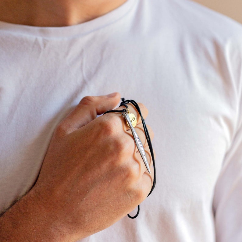 May include: A silver pendant necklace with a small round tag that says "118" and a long, thin, silver pendant with the word "SURVIVE" engraved on it. The necklace is on a black cord.