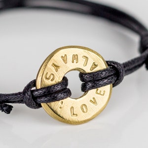 Op de afbeelding: Een armband van zwart koord met een ronde hanger van verguld metaal met de tekst "ALWAYS LOVE".