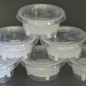 May include: A stack of six clear plastic containers with lids. Each container is filled with small, white, spherical beads. The containers are round with a slightly flared base and a snap-on lid. The background is a dark gray.