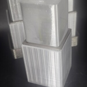 May include: A stack of several light gray, 3D-printed, square boxes. The top box has a cross-shaped indentation on its top surface. The lower boxes have vertical lines on their sides. The boxes are on a dark surface.