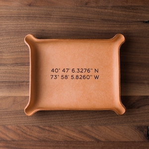 May include: A light brown leather tray with rounded edges. The tray has the coordinates "40° 47' 6.3276" N 73° 58' 5.8260" W" engraved on the surface.