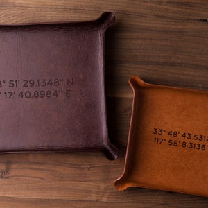 May include: Two leather trays with coordinates engraved on them. The top tray is brown and the bottom tray is tan. The coordinates on the top tray are 48° 51′ 29.1348" N 2° 17' 40.8984" E. The coordinates on the bottom tray are 33° 48' 43.5312" N 117° 55' 8.3136" W.