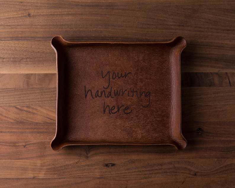 May include: A rectangular brown leather valet tray with raised, pinched corners. The center of the tray features the black engraved text "your handwriting here" in a casual, handwritten script style across three lines.