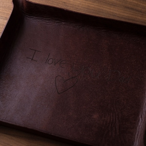 May include: A dark brown rectangular leather valet tray with raised edges. The center of the tray features an engraved inscription that reads I love you Dad, accompanied by a hand-drawn heart shape etched into the surface of the material.