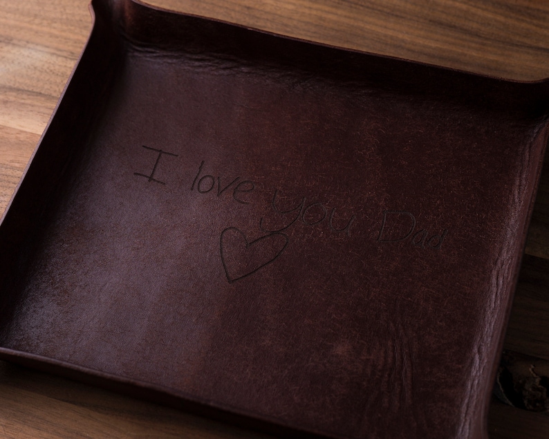 May include: A dark brown rectangular leather valet tray with raised edges. The center of the tray features an engraved inscription that reads I love you Dad, accompanied by a hand-drawn heart shape etched into the surface of the material.