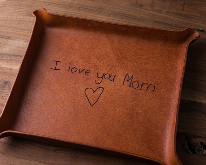 May include: A square brown leather valet tray with raised, folded corners. The center of the tray features the black engraved text "I love you Mom" positioned above a simple hand-drawn heart outline, creating a clear and legible message.