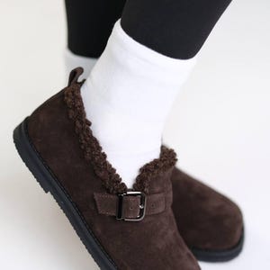 Pode incluir: Sapatos de camurça castanhos com fecho de fivela e acabamento em pele sintética. Os sapatos têm sola preta e são usados com meias brancas e leggings pretas. Os sapatos são um estilo slip-on.