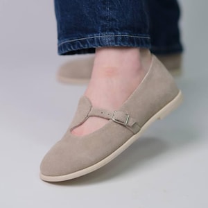 Könnte beinhalten: Beige Wildleder Mary Jane Ballerinas mit Schnallenverschluss. Die Schuhe haben eine runde Spitze und einen kleinen Riemen über dem Fuß. Die Schuhe werden mit blauen Jeans kombiniert.