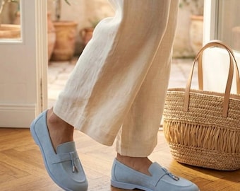 May include: A pair of light blue suede loafers featuring a tassel detail on the vamp, paired with beige linen trousers and a woven straw tote bag with tan leather handles, showcasing a classic slip-on design with a flat sole.