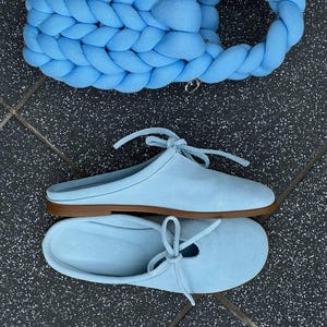 May include: A light blue handbag featuring a thick, braided fabric construction with a matching integrated handle, paired with a pair of light blue suede slip-on mules that have a thin tie detail across the top and a flat brown sole.