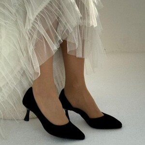 May include: A pair of black pointed-toe pumps with a low kitten heel, paired with a layered white tulle skirt featuring multiple tiers of sheer, ruffled fabric that create a voluminous and textured appearance around the lower legs.