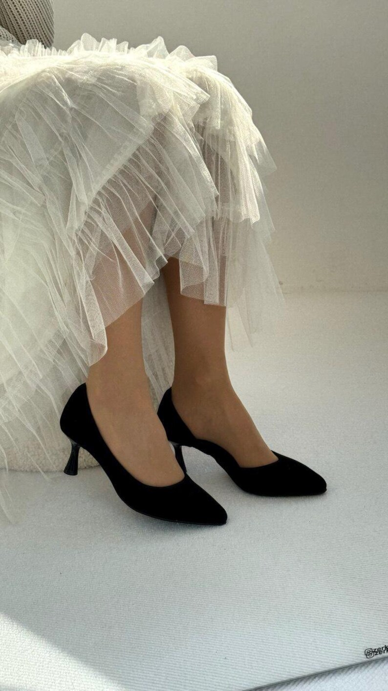 May include: A pair of black pointed-toe pumps with a low kitten heel, paired with a layered white tulle skirt featuring multiple tiers of sheer, ruffled fabric that create a voluminous and textured appearance around the lower legs.