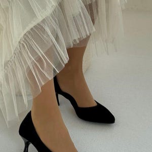 May include: A pair of black pointed-toe pumps with a slim kitten heel, paired with a layered off-white tulle skirt featuring multiple tiers of sheer, pleated fabric that falls in soft, voluminous ruffles around the lower legs and ankles.