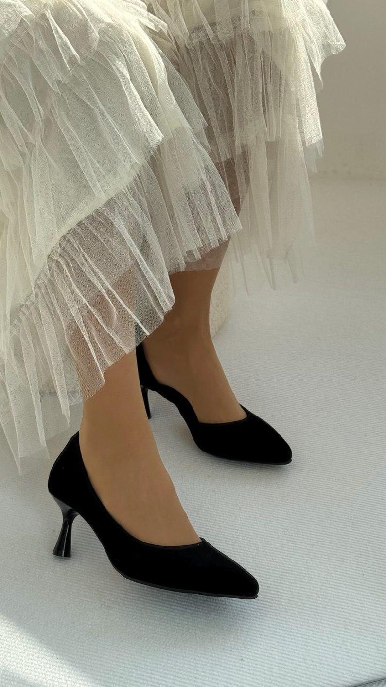 May include: A pair of black pointed-toe pumps with a slim kitten heel, paired with a layered off-white tulle skirt featuring multiple tiers of sheer, pleated fabric that falls in soft, voluminous ruffles around the lower legs and ankles.