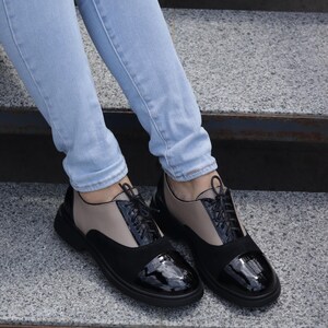 Puede incluir: Zapatos Oxford negros y beige con cordones negros y puntera brillante. Los zapatos tienen una suela negra gruesa y se usan con vaqueros azul claro. Una elecci&oacute;n de calzado elegante y moderno.