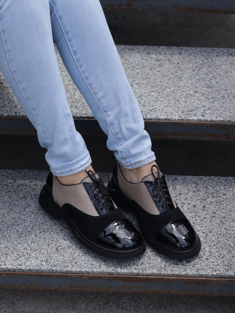 Puede incluir: Zapatos Oxford negros y beige con cordones negros y puntera brillante. Los zapatos tienen una suela negra gruesa y se usan con vaqueros azul claro. Una elecci&oacute;n de calzado elegante y moderno.