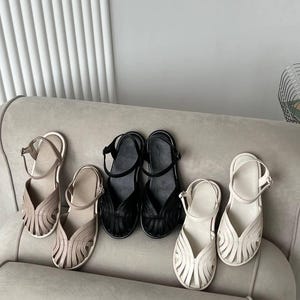 May include: Three pairs of closed-toe sandals in beige, black, and white. Each sandal features a woven, cage-like upper design with multiple thin straps, a rounded toe, and an adjustable ankle strap with a small metal buckle closure.