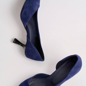May include: A pair of deep blue suede pumps featuring a pointed toe, a d'Orsay silhouette with an open side, and a slim, black flared kitten heel. The interior of the shoe is lined in a matching dark blue material with a visible brand logo.