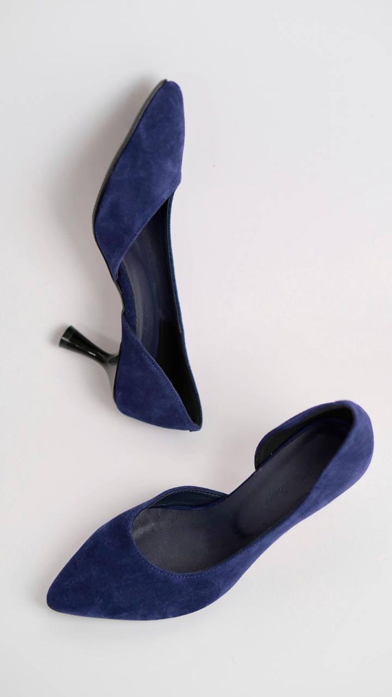 May include: A pair of deep blue suede pumps featuring a pointed toe, a d'Orsay silhouette with an open side, and a slim, black flared kitten heel. The interior of the shoe is lined in a matching dark blue material with a visible brand logo.