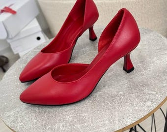 Puede incluir: Un par de zapatos de tacón rojo de punta con un detalle de corte en el lateral. Los zapatos están sobre una mesa redonda pequeña con un marco de alambre negro.