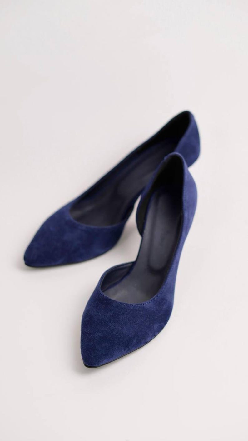 May include: A pair of deep blue suede pumps featuring a pointed toe and a d'Orsay silhouette with an open side design. The interior lining is dark, and the shoes have a slim heel profile, showcasing a solid color finish throughout the exterior.