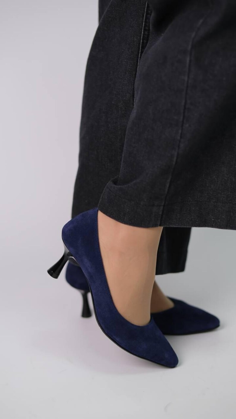 May include: A pair of navy blue suede pumps featuring a pointed toe and a black kitten heel. The shoes are worn with dark charcoal grey denim trousers that fall just above the ankle, showcasing the sleek silhouette of the footwear.