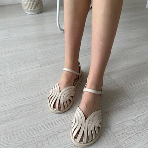 May include: A pair of off-white leather sandals featuring a cage-style upper with multiple curved straps, a closed rounded toe, and an adjustable ankle strap with a small metal buckle. The sandals have a flat sole and a neutral, light-colored finish.