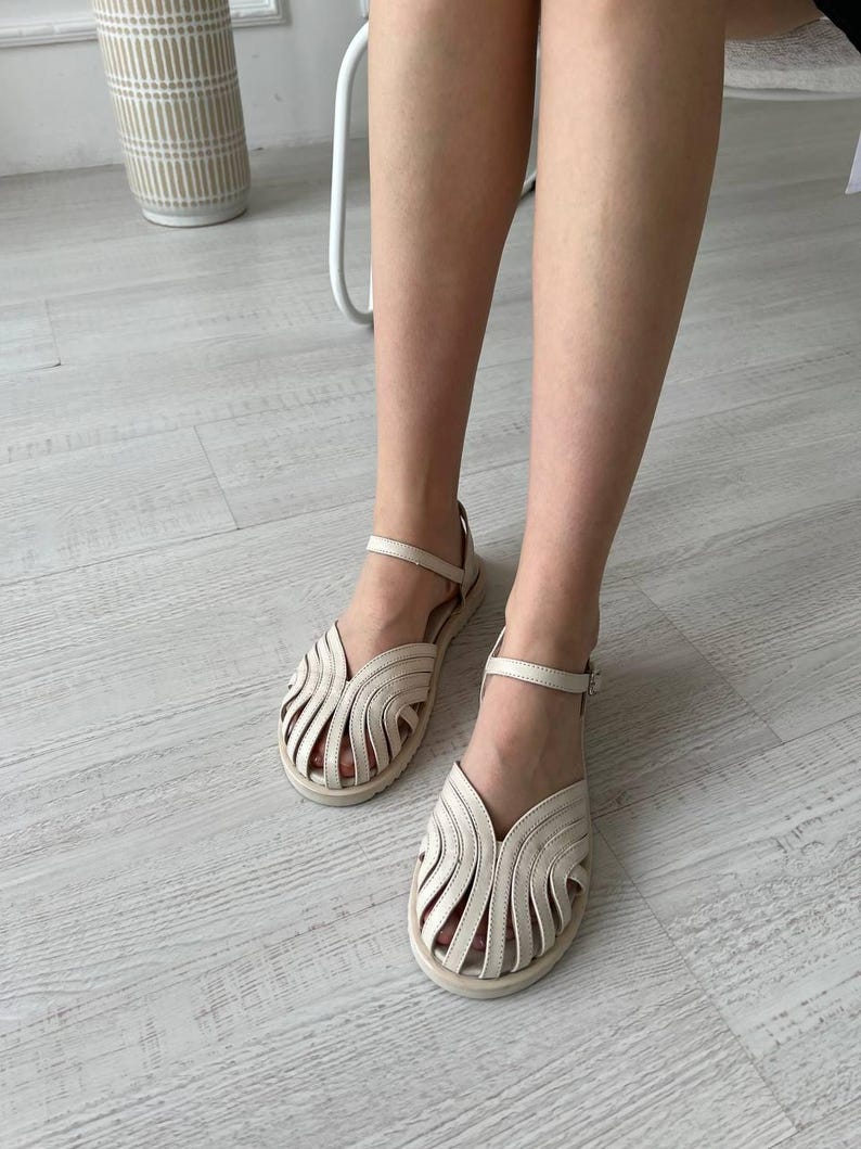 May include: A pair of off-white leather sandals featuring a cage-style upper with multiple curved straps, a closed rounded toe, and an adjustable ankle strap with a small metal buckle. The sandals have a flat sole and a neutral, light-colored finish.