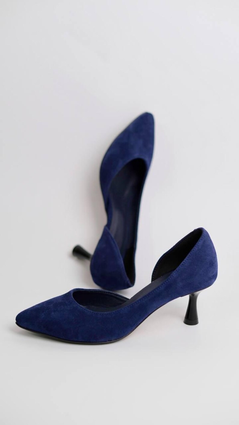 May include: A pair of deep blue suede pumps featuring a pointed toe, a d'Orsay silhouette with a side cutout, and a short, black kitten heel. The shoes have a smooth, matte texture and a clean, structured design with a dark interior lining.