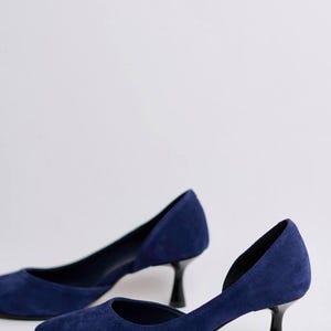 May include: A pair of navy blue suede pumps featuring a pointed toe, a low-cut side profile, and a black kitten heel. The shoes have a smooth, matte texture and a clean, simple design with a closed heel counter and a structured, narrow silhouette.