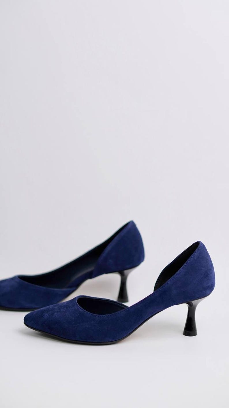 May include: A pair of navy blue suede pumps featuring a pointed toe, a low-cut side profile, and a black kitten heel. The shoes have a smooth, matte texture and a clean, simple design with a closed heel counter and a structured, narrow silhouette.