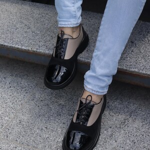 Puede incluir: Zapatos Oxford bicolor con punteras de charol negro y paneles de ante negro. Los zapatos tienen la parte superior de cuero beige y cordones negros. Llevados con vaqueros azul claro.