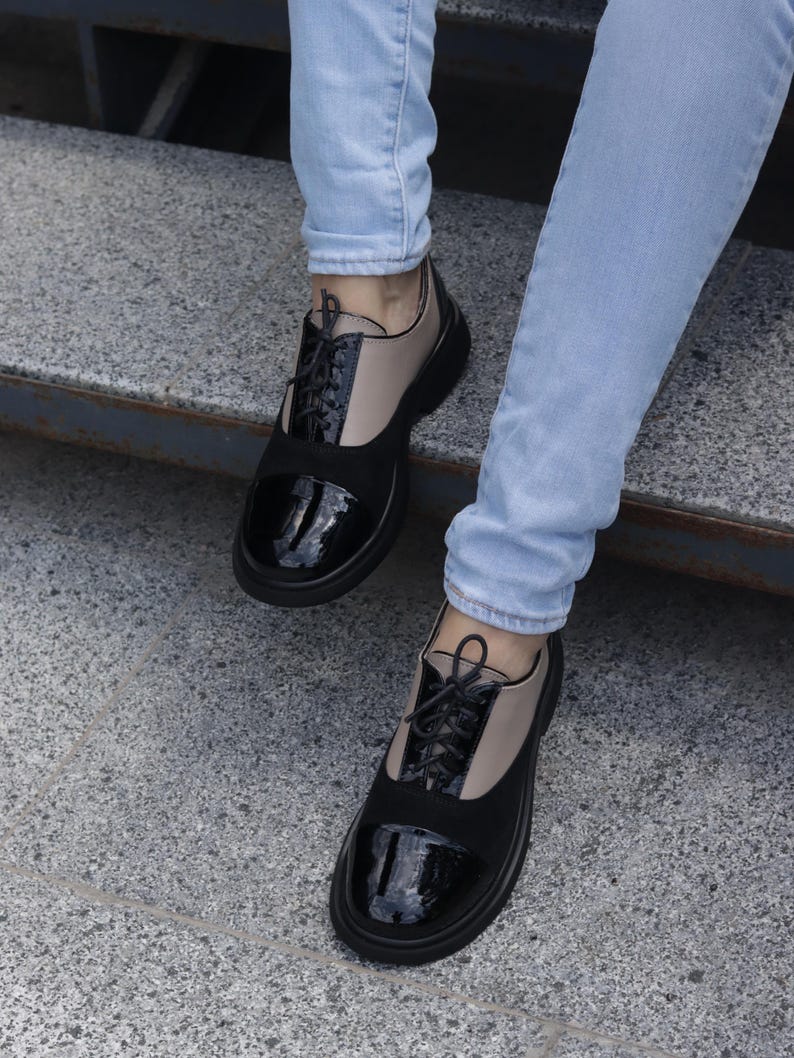 Puede incluir: Zapatos Oxford bicolor con punteras de charol negro y paneles de ante negro. Los zapatos tienen la parte superior de cuero beige y cordones negros. Llevados con vaqueros azul claro.