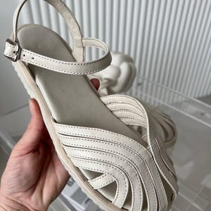 May include: A cream-colored sandal featuring a closed-toe design with multiple overlapping leather straps, a flat sole, and an adjustable ankle strap with a silver-toned metal buckle. The upper construction consists of curved, stitched leather strips.
