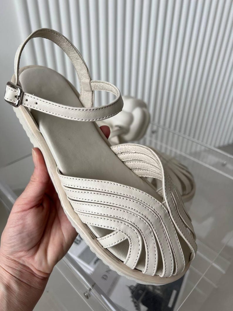 May include: A cream-colored sandal featuring a closed-toe design with multiple overlapping leather straps, a flat sole, and an adjustable ankle strap with a silver-toned metal buckle. The upper construction consists of curved, stitched leather strips.
