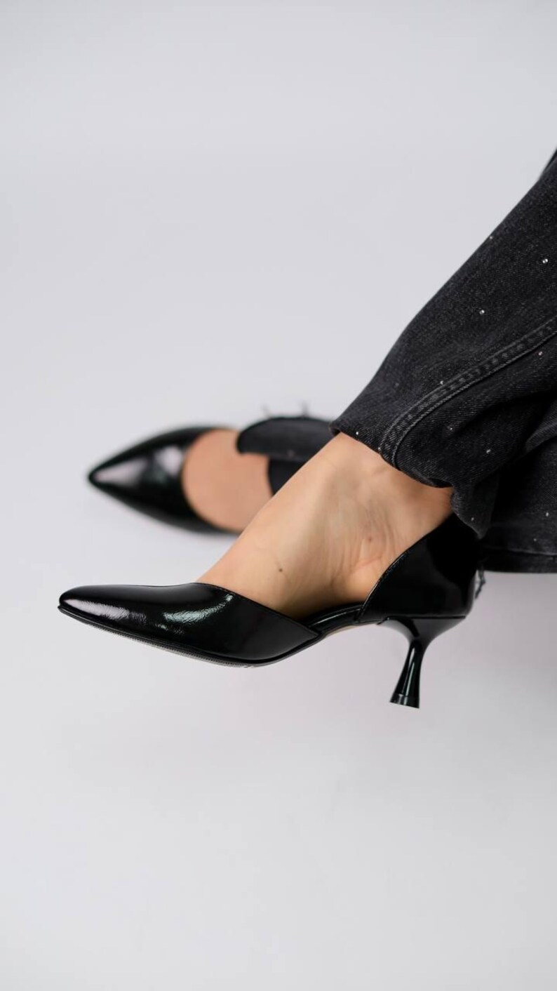 May include: A pair of black patent leather d'Orsay pumps featuring a pointed toe and a low kitten heel. The shoes have a glossy finish and a cut-out side design, paired with dark grey denim jeans that have subtle silver embellishments.