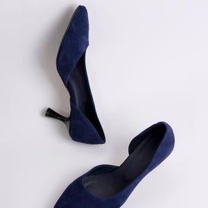 May include: A pair of navy blue suede pumps featuring a pointed toe, a d'Orsay silhouette with an open side, and a slim black kitten heel. The shoes have a smooth, matte texture and a clean, minimalist design with a deep blue color throughout.