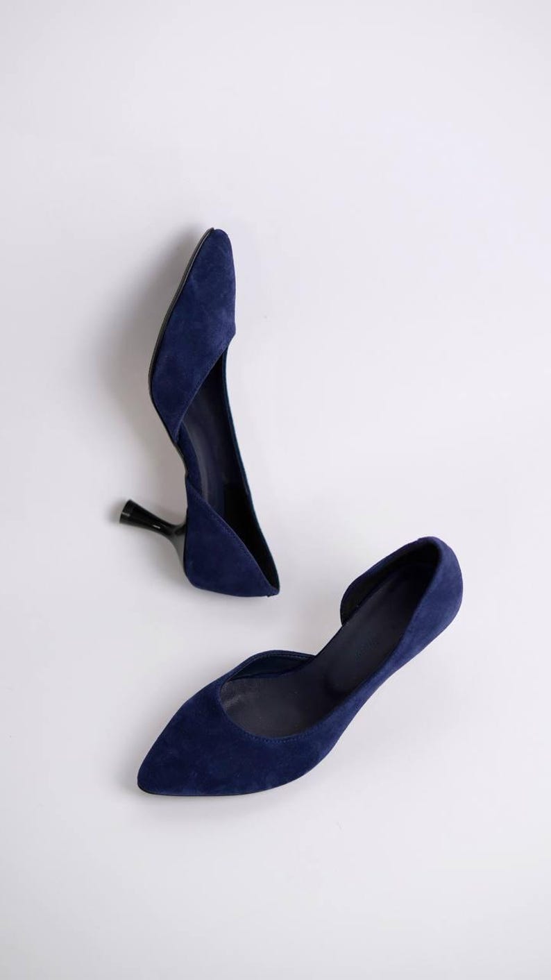May include: A pair of navy blue suede pumps featuring a pointed toe, a d'Orsay silhouette with an open side, and a slim black kitten heel. The shoes have a smooth, matte texture and a clean, minimalist design with a deep blue color throughout.