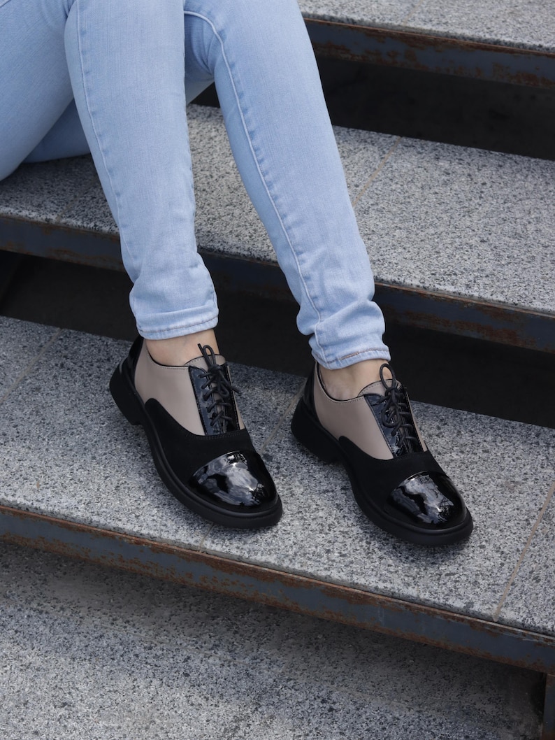 Puede incluir: Zapatos Oxford negros y beige con puntera de charol brillante y cordones negros. Los zapatos tienen una suela negra gruesa. Los zapatos se llevan con vaqueros azul claro. Sobre una superficie de hormig&oacute;n gris.