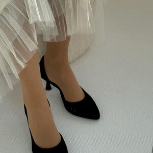 May include: A pair of black pointed-toe pumps with a low kitten heel, featuring a smooth finish and a closed-toe design. The shoes are worn with a white layered tulle skirt that falls above the ankles, creating a contrast in texture and color.