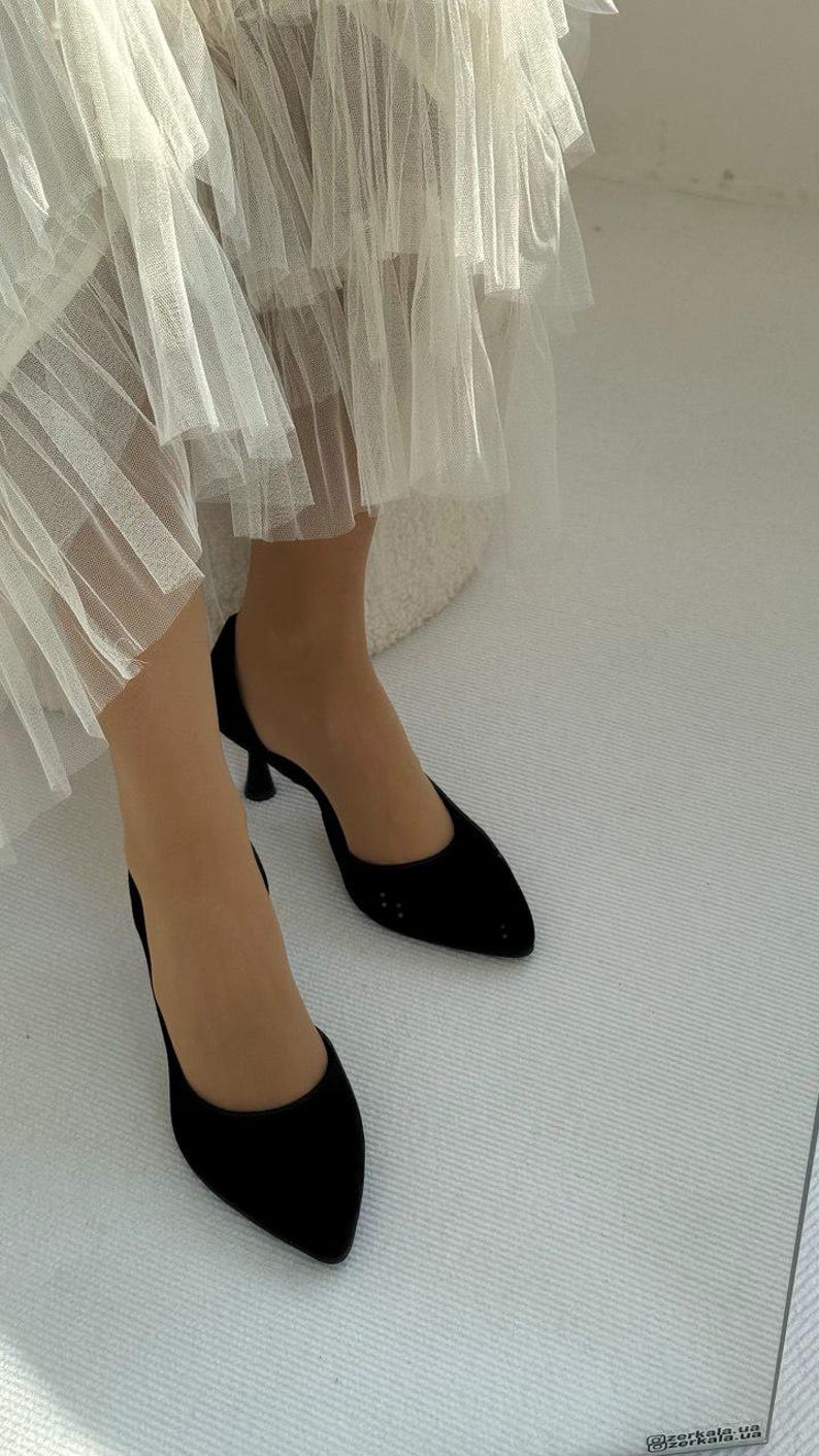 May include: A pair of black pointed-toe pumps with a low kitten heel, featuring a smooth finish and a closed-toe design. The shoes are worn with a white layered tulle skirt that falls above the ankles, creating a contrast in texture and color.