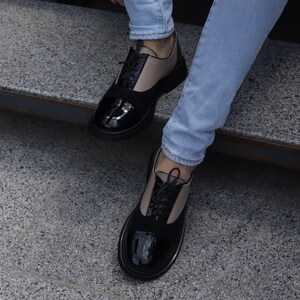 Puede incluir: Zapatos Oxford negros y beige con puntera de charol brillante y material similar al ante en la parte superior. Los zapatos tienen cordones negros y una suela gruesa negra. La persona lleva vaqueros azul claro.
